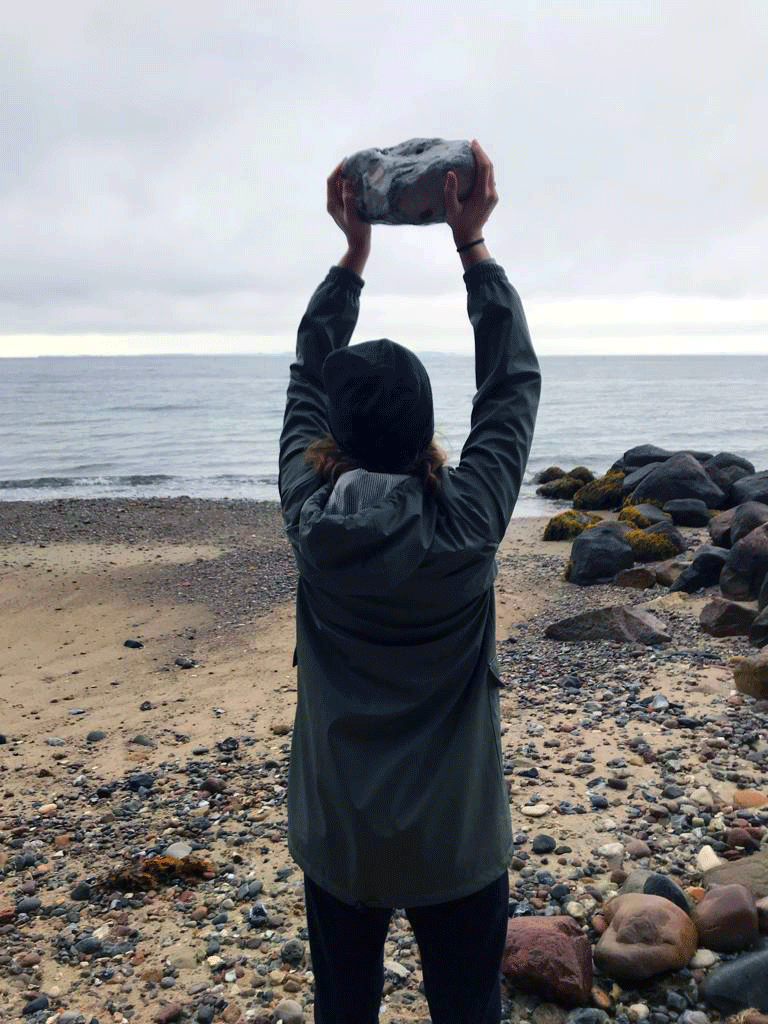 Eine Frau steht mit dem Rücken zur Kamera und reckt am Meer einen großen Stein in den Himmel.